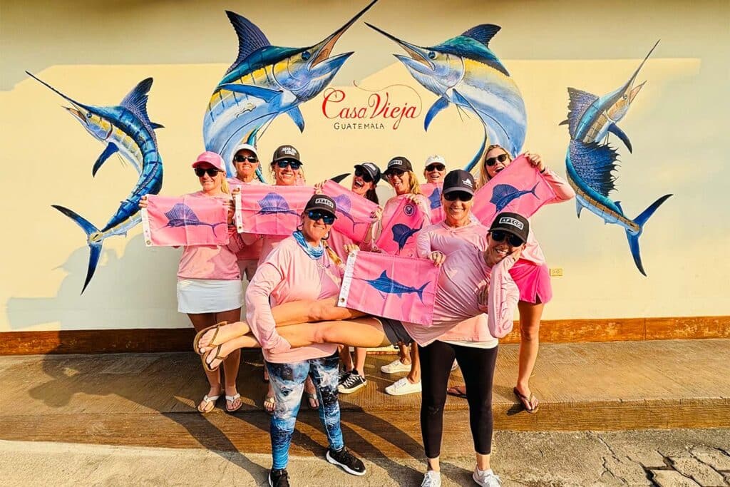 A group of women wearing pink shirts and holding pink marlin release flags stand in front of the Casa Vieja sign.