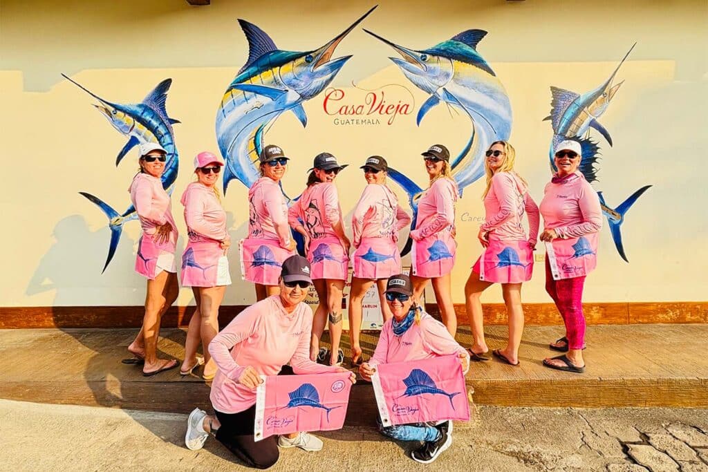 A group of women wearing pink shirts and holding pink marlin release flags stand in front of the Casa Vieja sign.