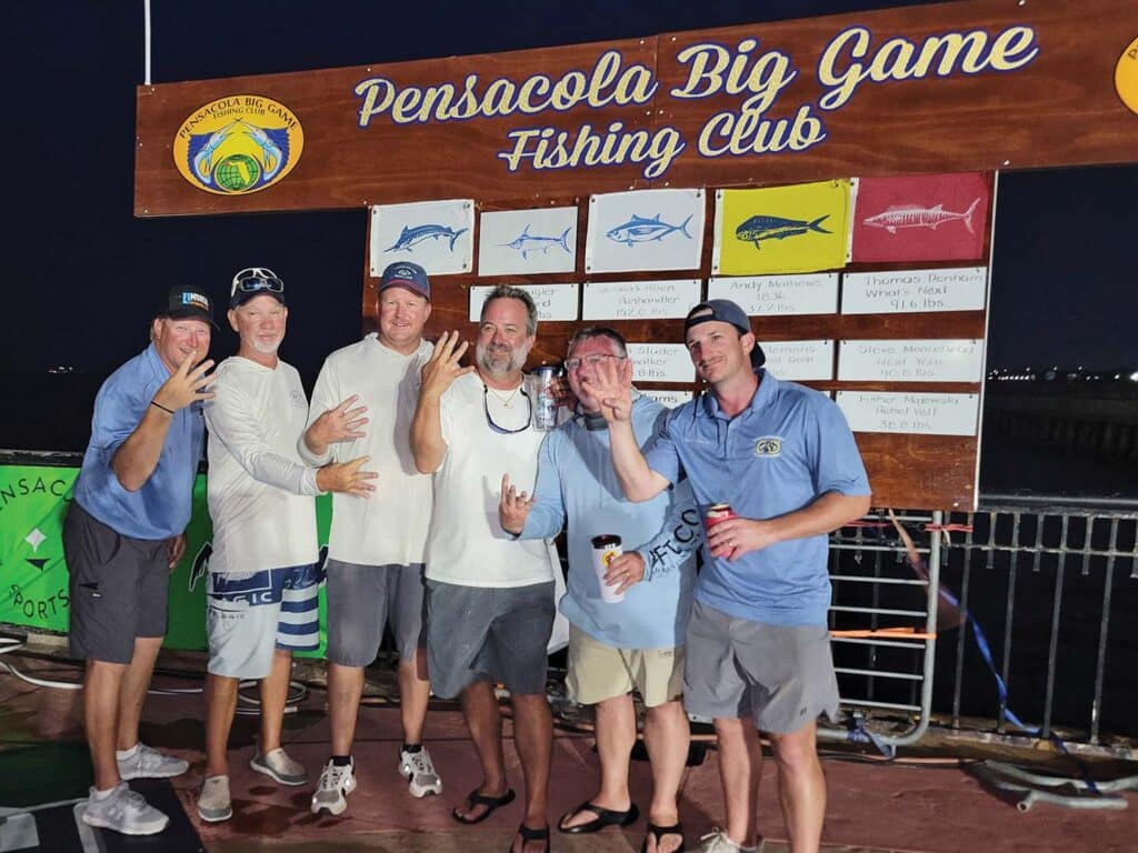 Six men stand in celebration. Behind them is a leaderboard covered in sport-fishing release flags. The sign is labeled Pensacola Big Game Fishing Club.