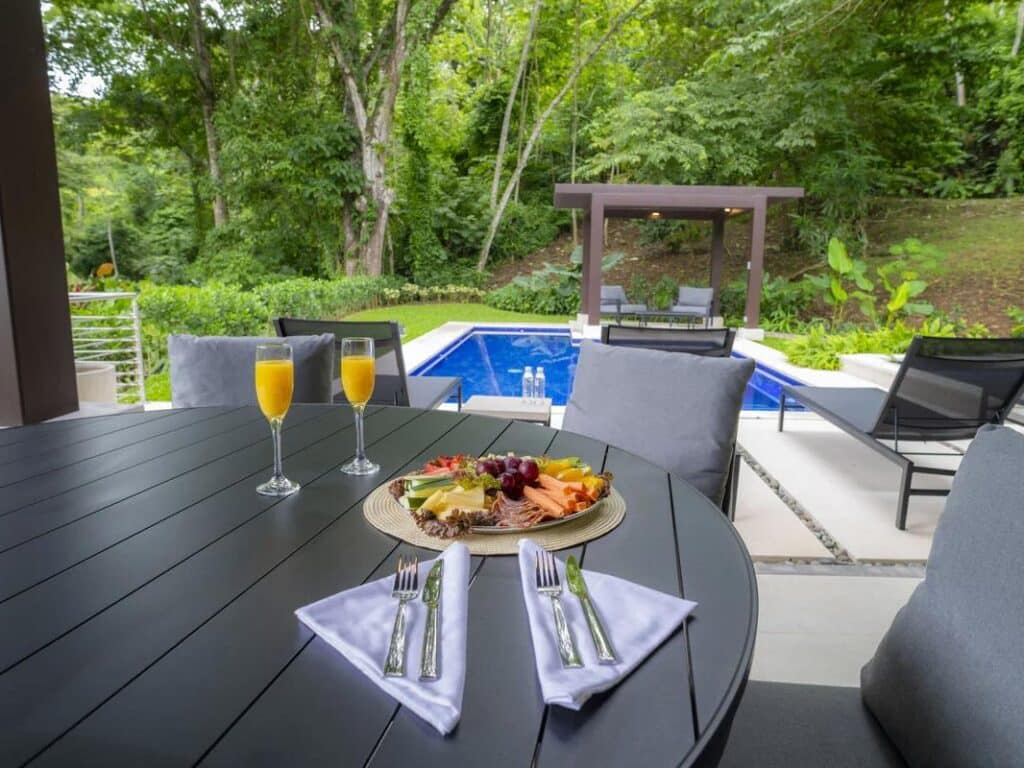 Outdoor patio with a view of a pool and trellis gazebo. On the patio table is two glasses of mimosa, and a charcuterie plate.