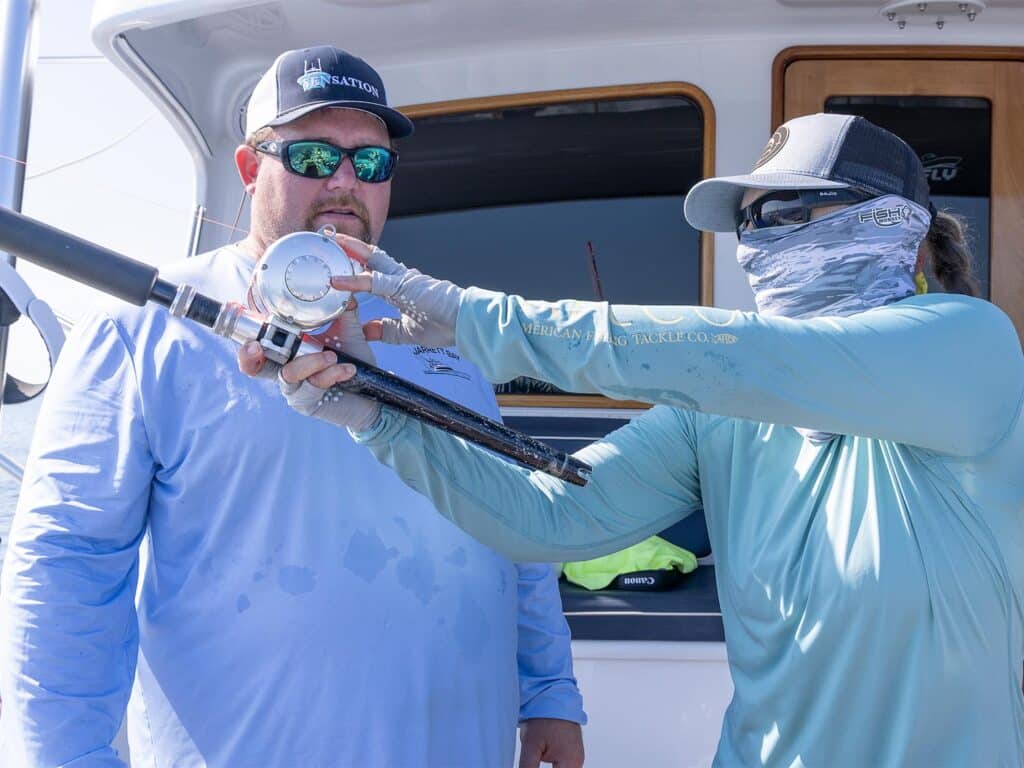 A woman holds up a fishing rod and reel to demonstrate practices to an Expedtions attendee. Both are wearing sun-protective eyewear and clothing.
