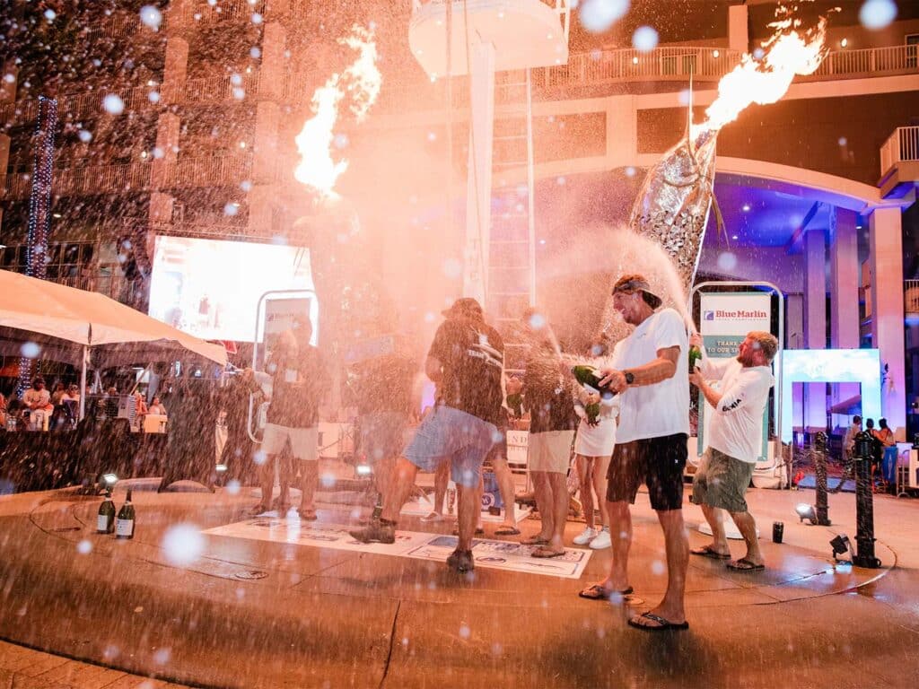 A sport-fishing team celebrates at an awards ceremony by popping bottles of champagne, spraying the liquid into the air. Fire spews from two metal marlin statues.