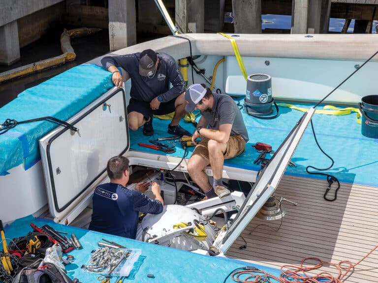 Three boat crew administering repairs to a sport-fishing boat via a hatch opening in the cockpit. protective tarps are laid over the transom and hardwood of the cockpit's flooring.