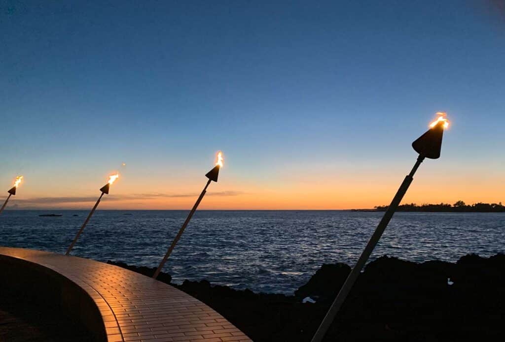Night-time view of lit torches on a railing. Ocean can be seen in the background with a sunset on the horizon.