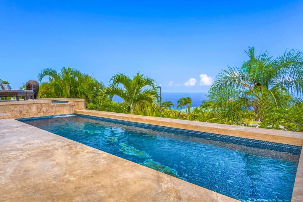 View of the pool at a Kona, Hawaii resort. Lush tropical trees and palms surround the concrete patio space into the horizon.