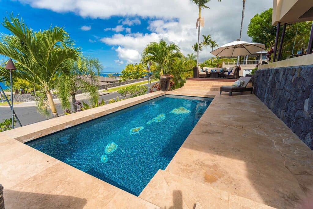 View of the pool at a Kona, Hawaii resort. Lush tropical trees and palms surround the concrete patio space into the horizon.