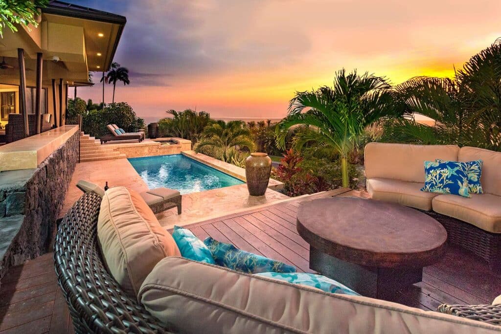 View of the sunset from the outdoor patio of a Hawaiian resort. The orange sky silhouettes trees, and is reflected in the patio's pool.