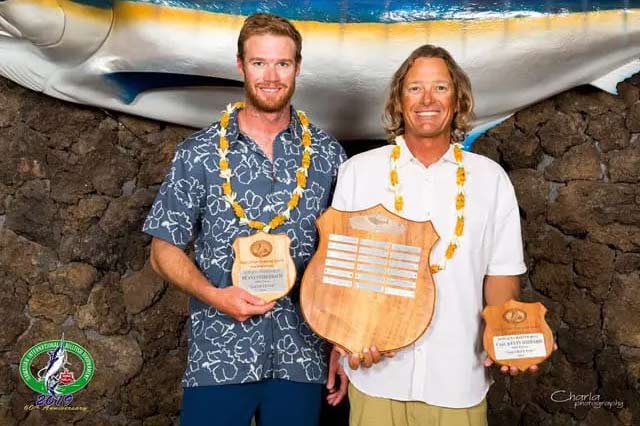 Two anglers stand side-by-side at an awards ceremony while holding plaques and trophies.
