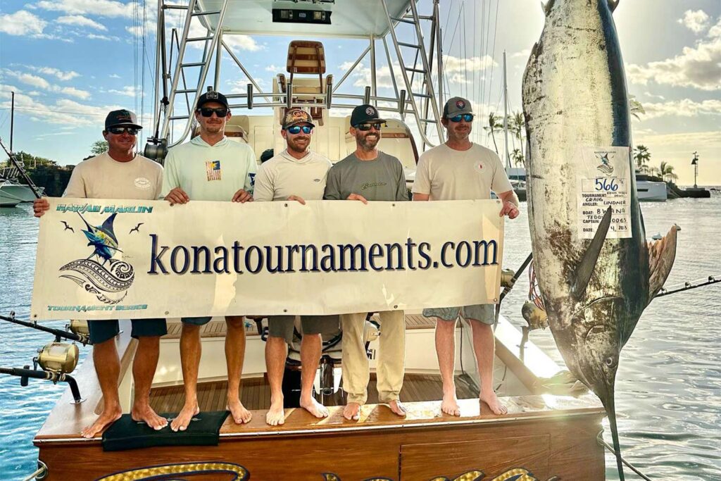 A sport-fishing crew stand on the transom of their sport-fishing boat next to a weighed marlin. They hold up a sign with "konatournaments.com" on it.