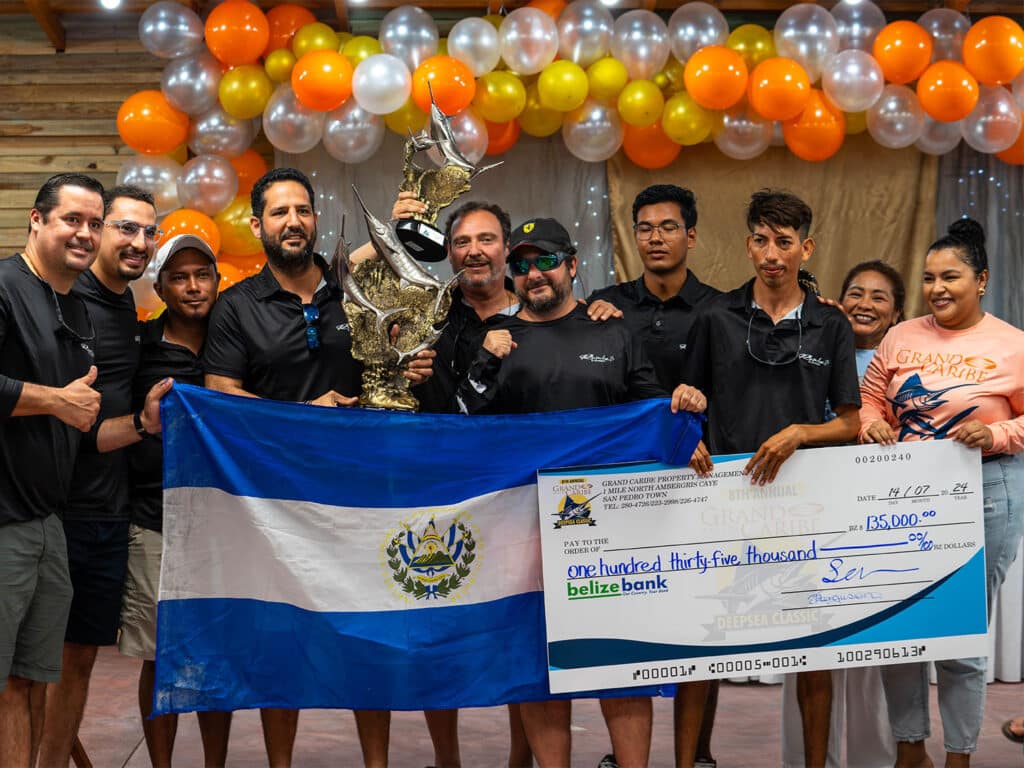 A team of anglers standing at an awards ceremony. Several are holding up large trophies, the flag of El Salvador, and an over-sized check.