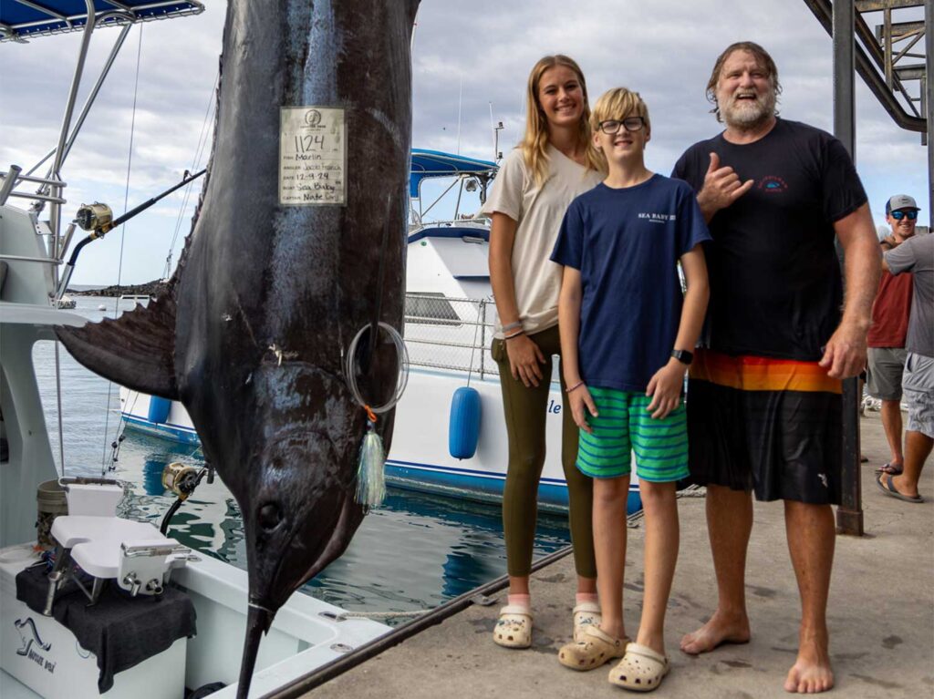 A fishing family stands next to a grander blue marlin being weighed dockside.