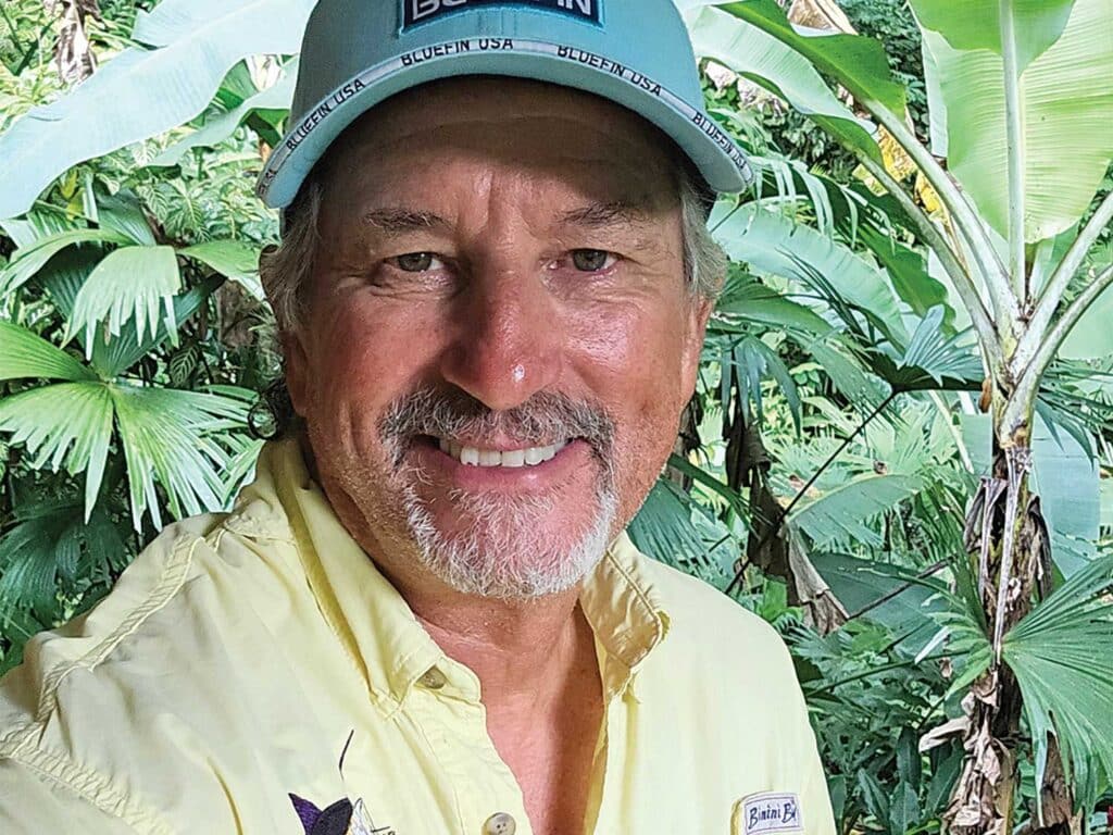 Bill Boyce smiles for the camera with a backdrop of lush tropical plants behind him.