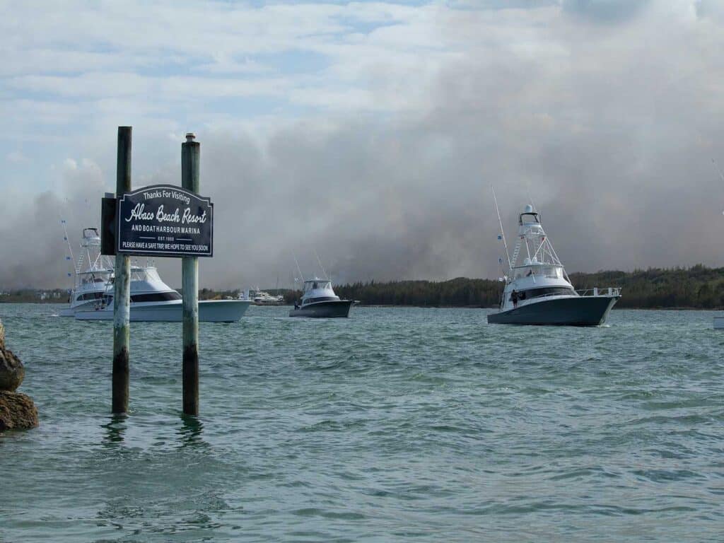 A fleet of sport-fishing boats in Marsh Harbor. A sign in the water reads "Thanks for visiting Abaco Beach Resort and Boat Harbor Marina. In the distances smoke from wildfires rises into the sky.