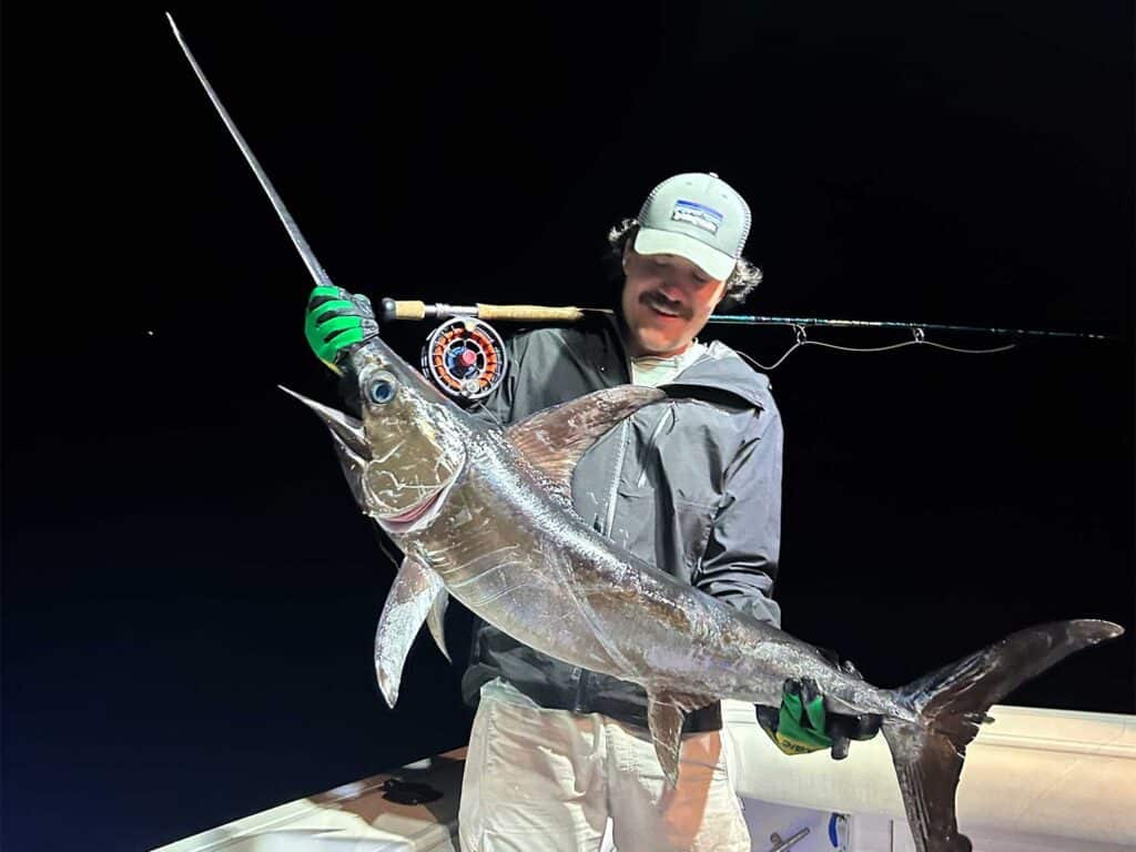 An angler stands in the cockpit of a sport-fishing boat and holds up a swordfish.