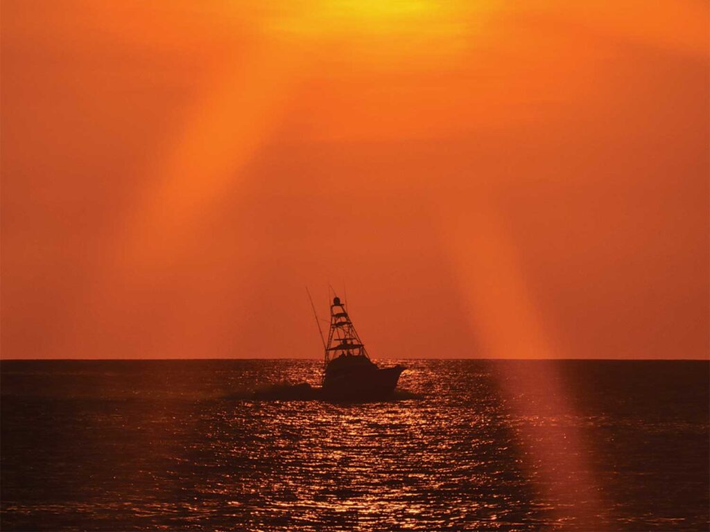 A sport-fishing boat silhouetted against the ocean horizon, lit up orange as the sun sets.