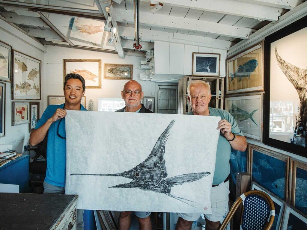 Three men hold up an illustration of a white marlin.