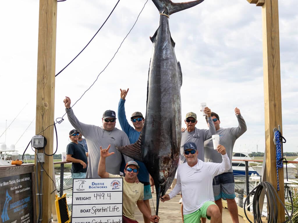 A team of anglers stand next to a large blue marlin being weighed at the docks.