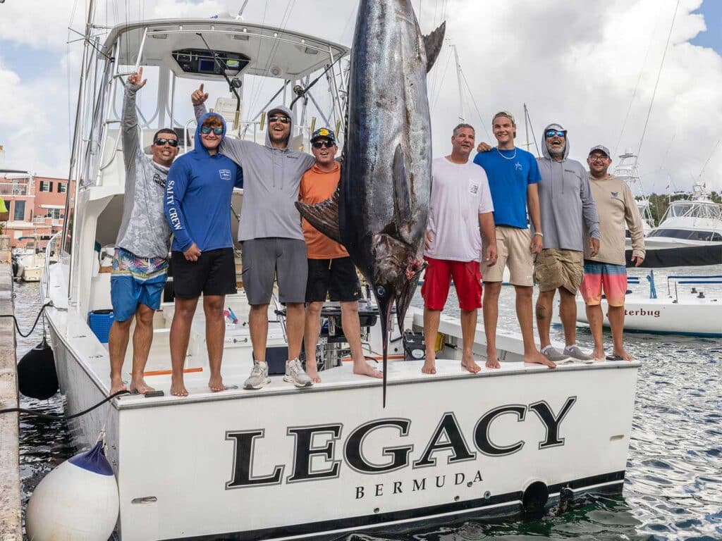 Team Legacy stands on the transom of their sport-fishing boat next to a large blue marlin being weighed.
