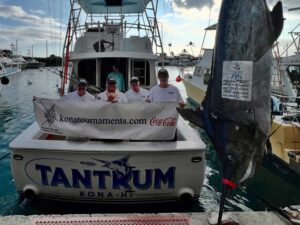 Team Tantrum stands in the cockpit of their sport-fishing boat next to a 700+ pound blue marlin being weighed dockside.