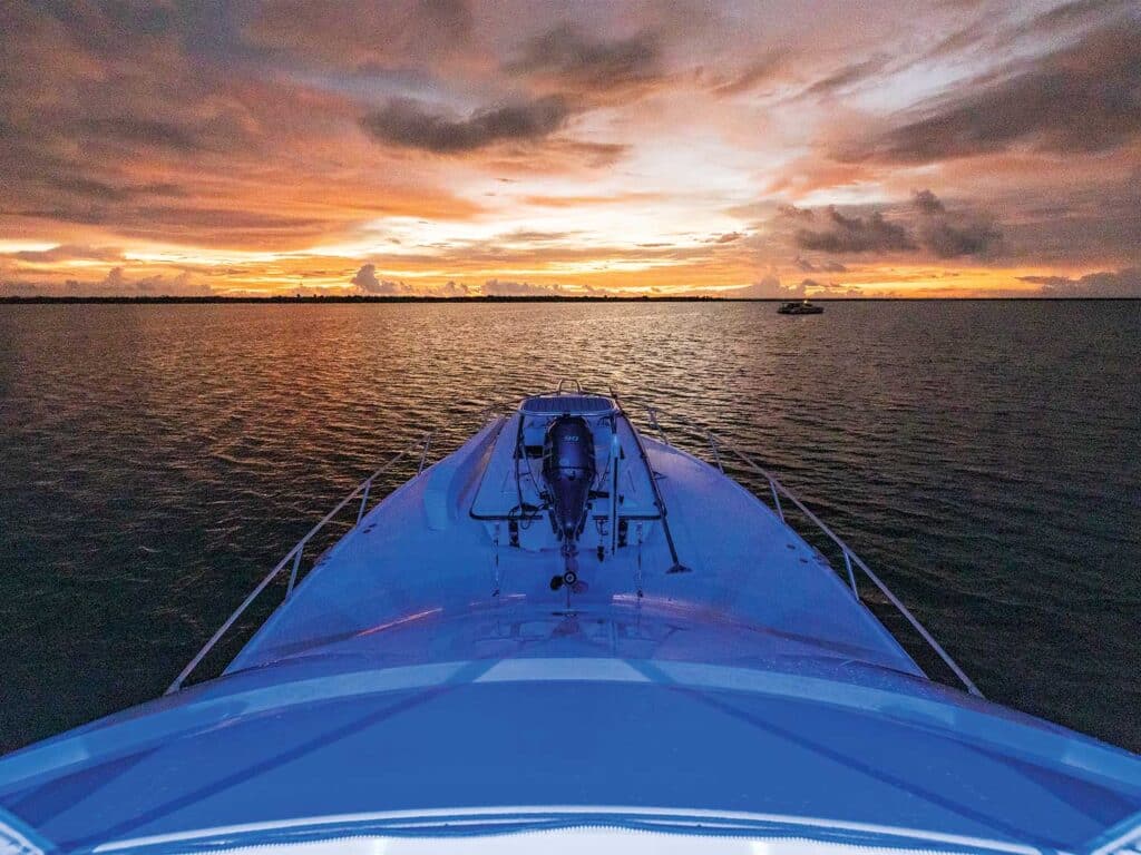 A view from the bow of a sport-fishing boat. A skiff sits securely mounted to the bow. In the horizon the sun sets against the ocean.